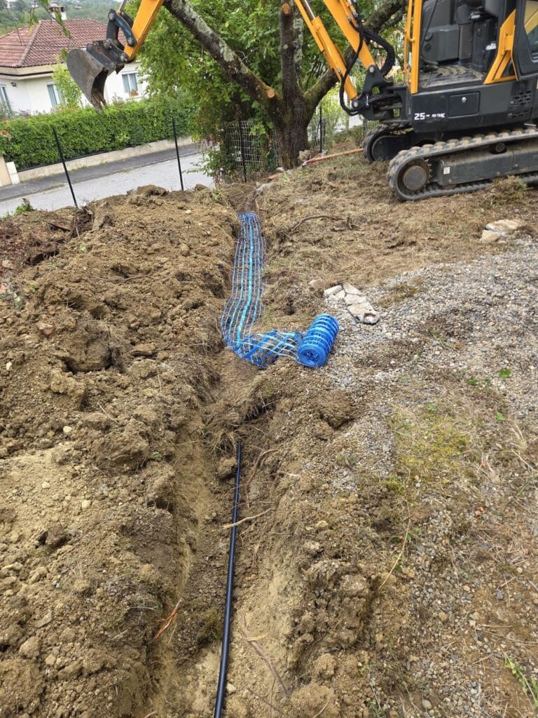 Mini-pelle 25-OK creuse une tranchée, posant une gaine de protection bleue pour câbles et une gaine noire. Travaux d'enfouissement.